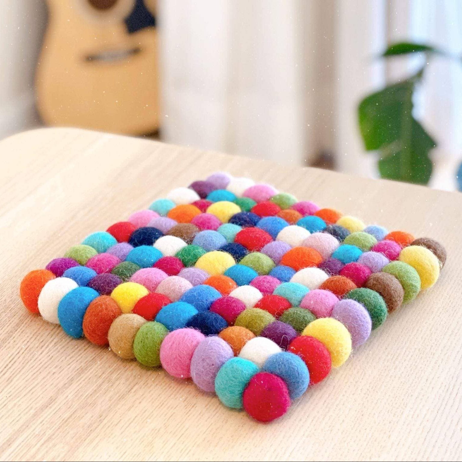 Colorful felt ball coaster on a wooden surface with a guitar and plant in the background